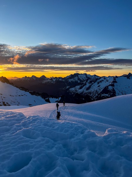 Climbing team of three, roped up on the inspiration glacier at sunrise with long clouds over several jagged peaks in the North Cascades.