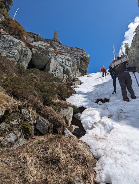 Two people carrying heavy packs using an ice axe each while climbing up a steep, snow filled gully.