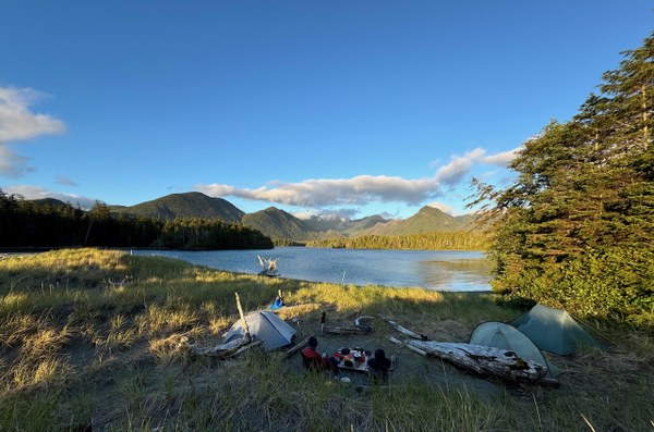 Landscap photo.  3 tents in the foreground, a body of water, mountains in the distance in evening sunlight